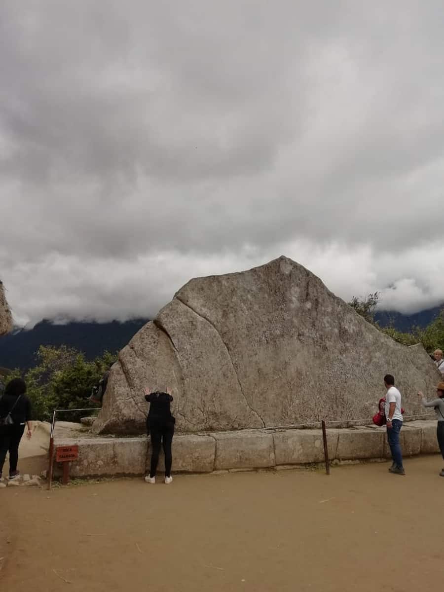 Triangular 15 foot rock with two people standing in front of it called Sacred Rock at Machu Picchu